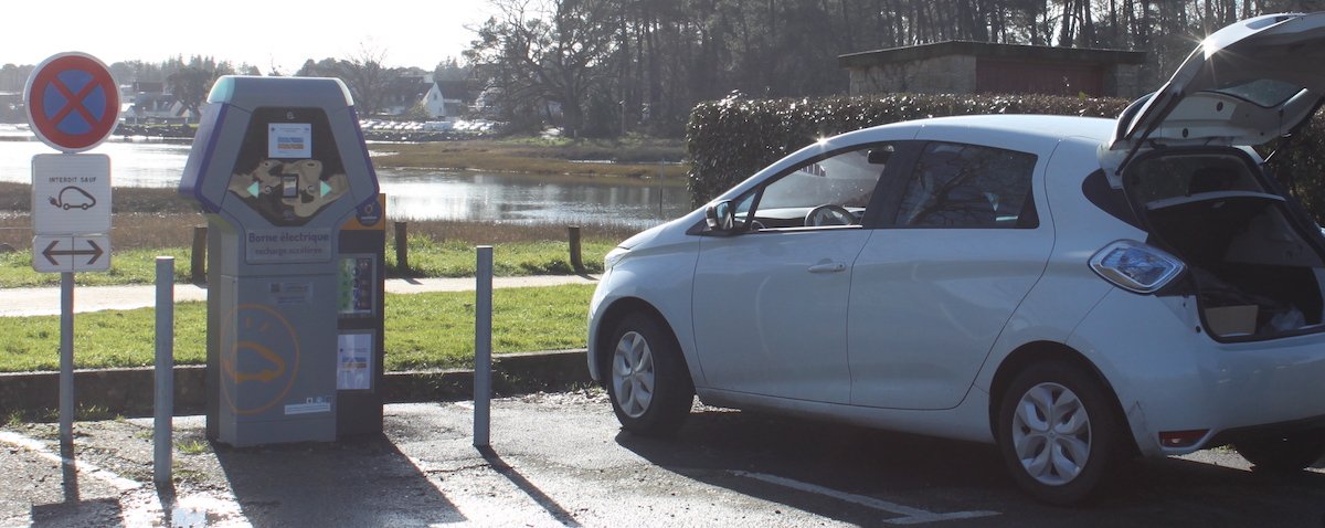 Photographie d'une borne de recharge électrique prise à Vannes à Conleau. Une voiture électrique blanche est sur la place prête à être rechargée.